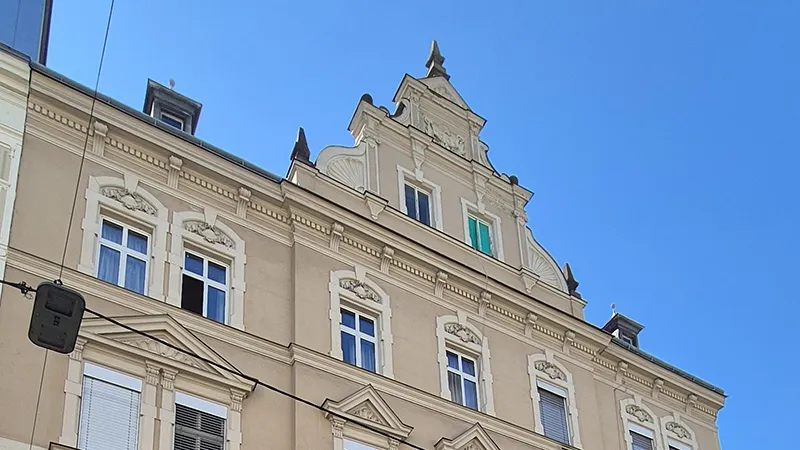 Ein kunstvolles, beiges Gebäude mit dekorativen weißen Verzierungen unter strahlend blauem Himmel. Zu den Besonderheiten zählen aufwendige Zierleisten und ein blaugrünes Fenster, das einen Kontrast zu den anderen Fenstern bildet.