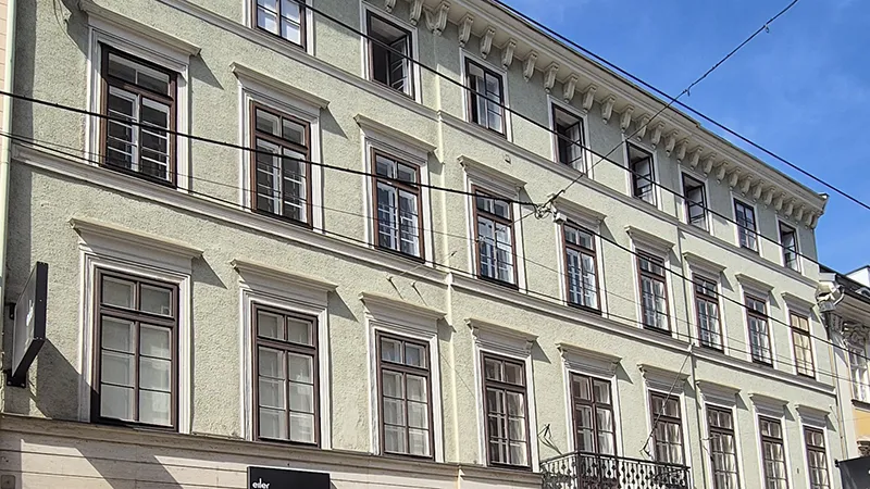Ein historisches vierstöckiges Gebäude mit hellgrüner Fassade, symmetrischen Fenstern und verzierten Gesimsen unter strahlend blauem Himmel. Die Szene wirkt ruhig und elegant.